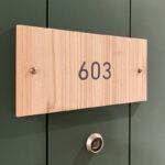 room number on wooden sign mounted on dark green door above peephole